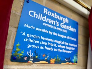 An aquatic-themed plaque on a brick wall which reads "Roxburgh Children's Garden. Opened 28 April 2026. Made possible by the League of Friends. "A garden becomes magical the moment children step into it, where laughter grows as freely as the flowers."
