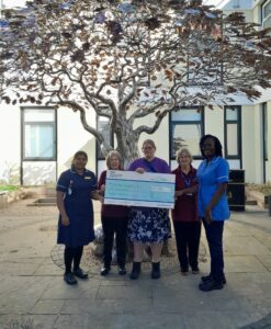 A photo of Maggie Drew holding a giant novelty cheque worth £30,000 to The Queen Elizabeth Hospital Charitable Fund, next to Cancer Ward Manager, a nurse and two volunteers.