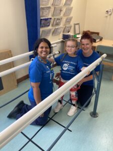 A photo of physiotherapists helping to support a young patient during their therapy sessions. They are all wearing "I'm raising money for QEHKL Charity" t shirts